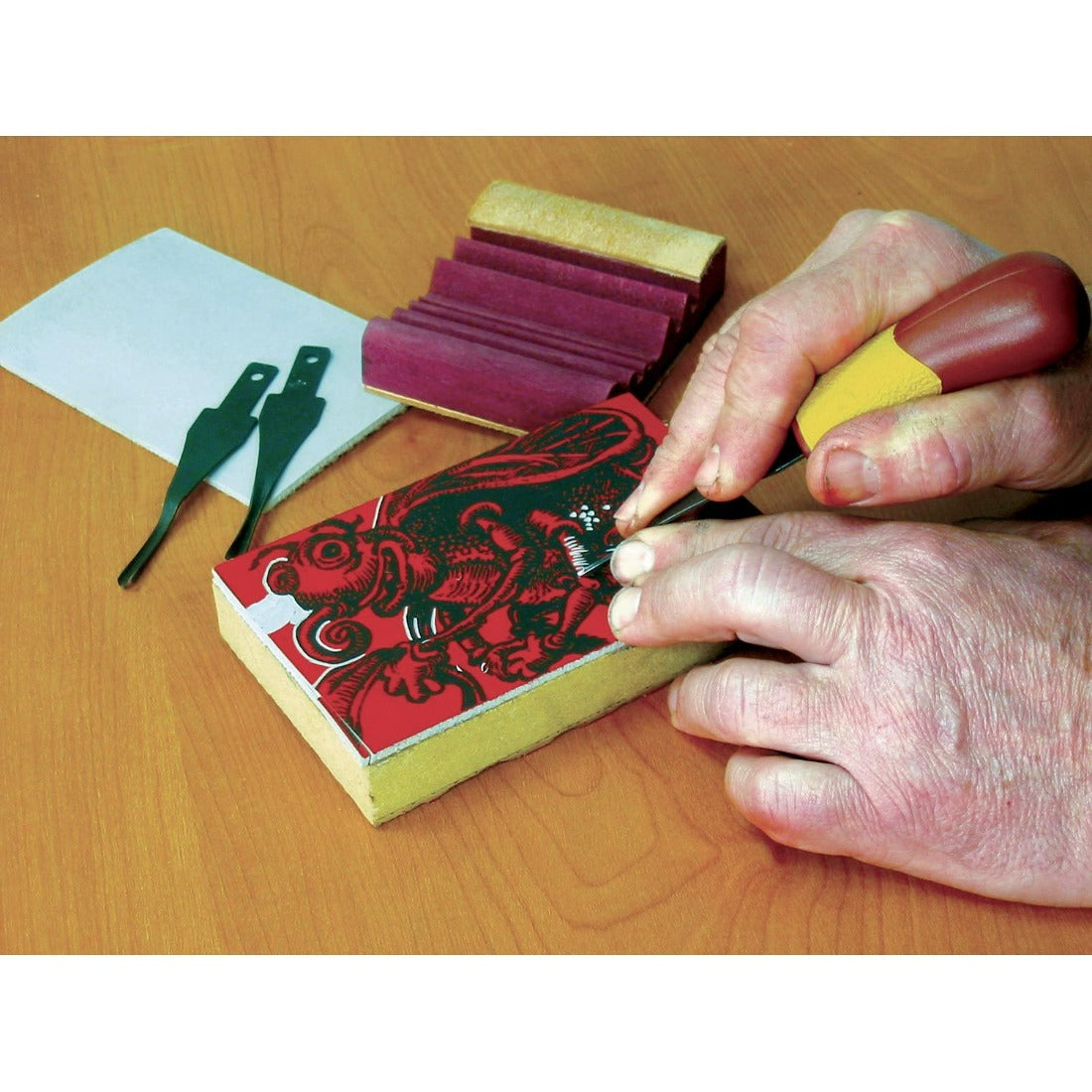image showing one wide gouge being used to carve into a lino which is affixed to a block. User carving aa relief image into the lino. A slipstrop and to other gouge blades lying on the table