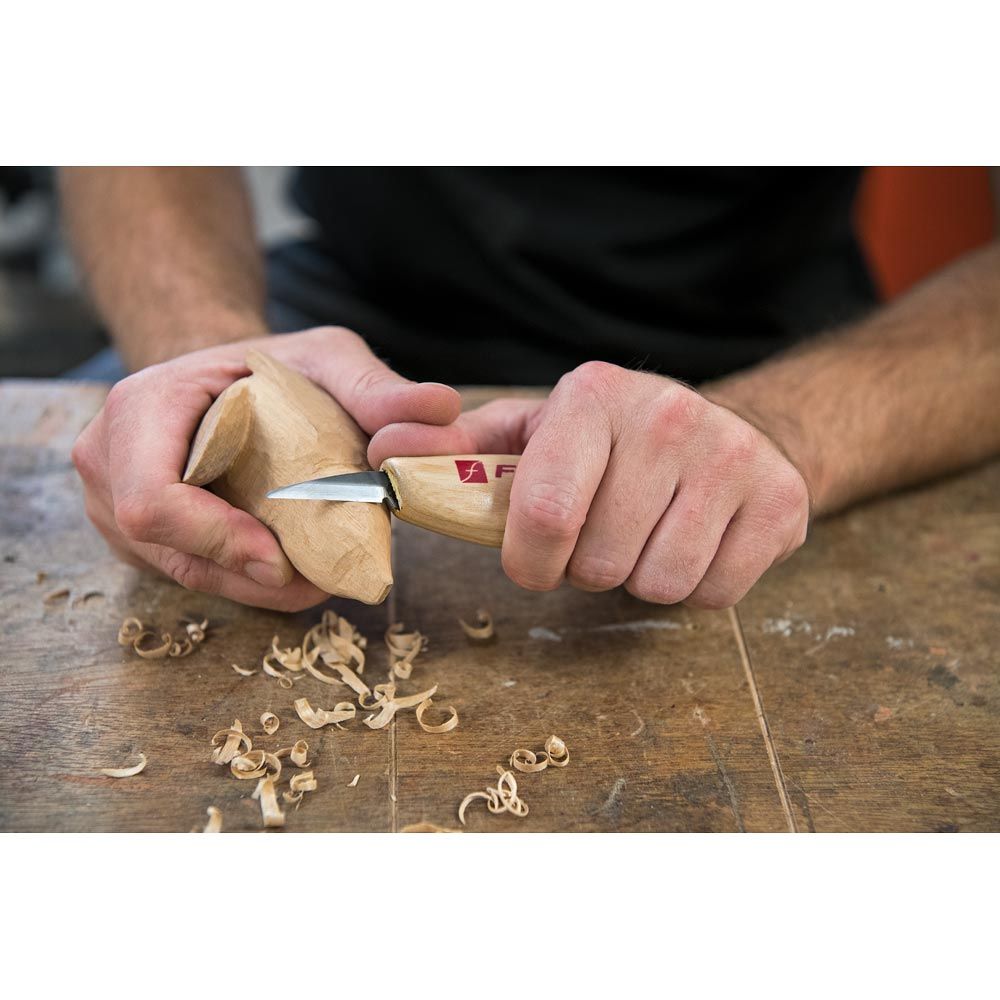 picture of a carver, carving a wooden figure on a bench. Shavings shown on the bench from the carving.